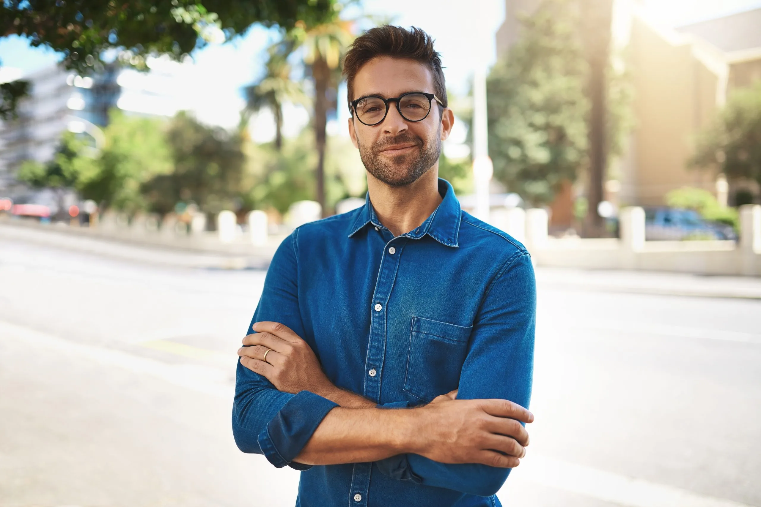Man in denim shirt with arms crossed standing outdoors on a city street