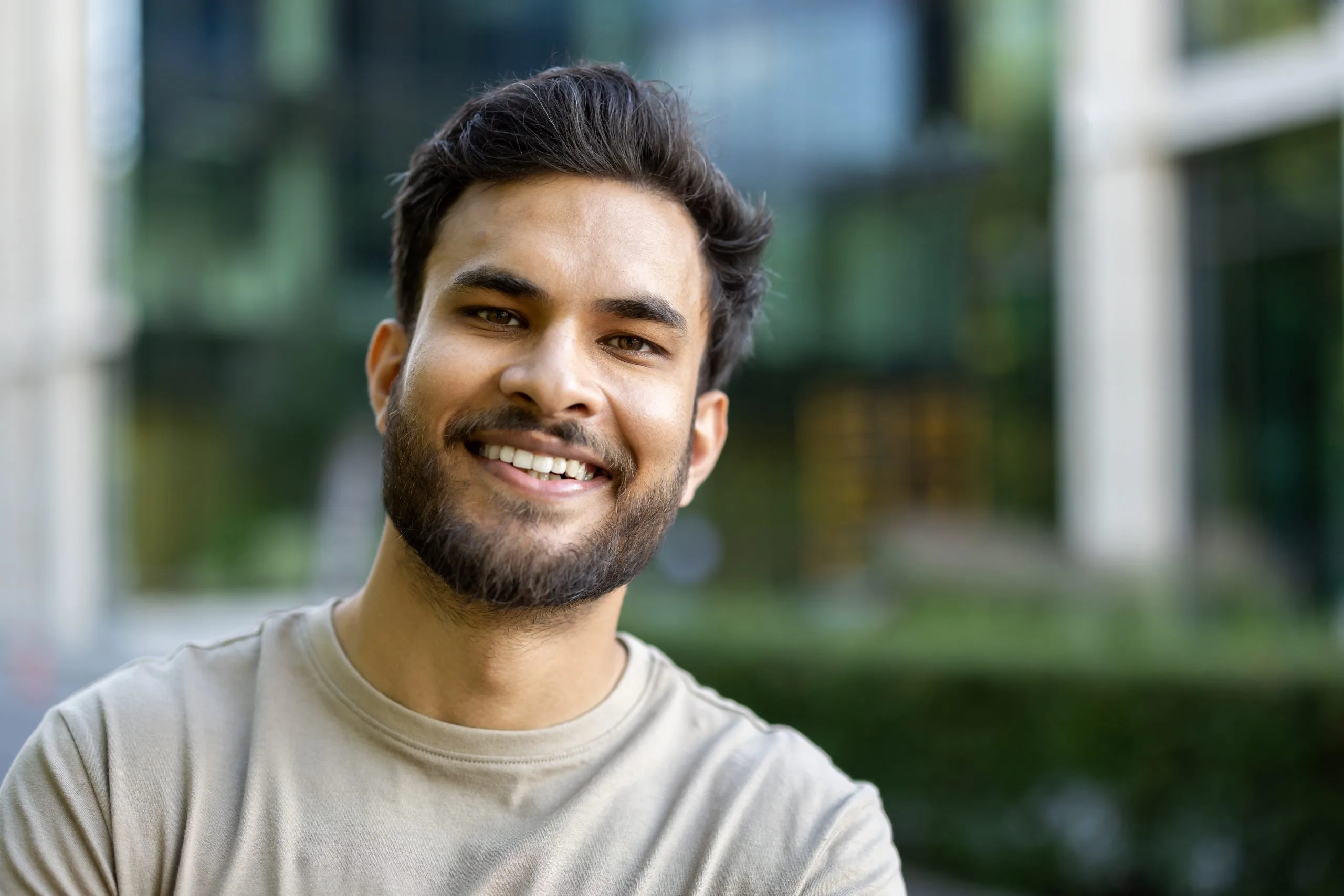 Smiling man with beard in gray shirt outdoors with blurred greenery behind