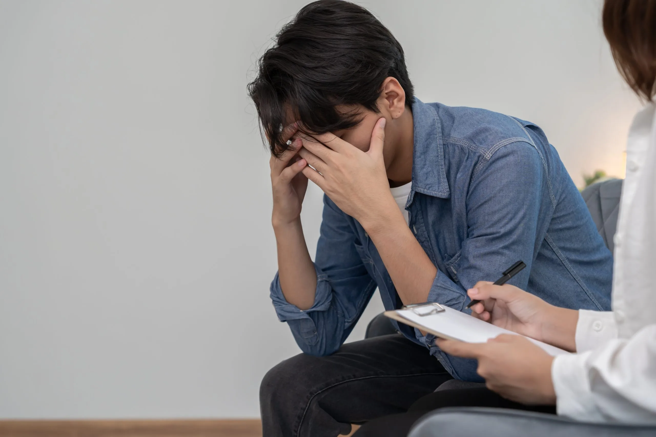 Woman covering face with hands in distress during consultation with healthcare provider holding clipboard