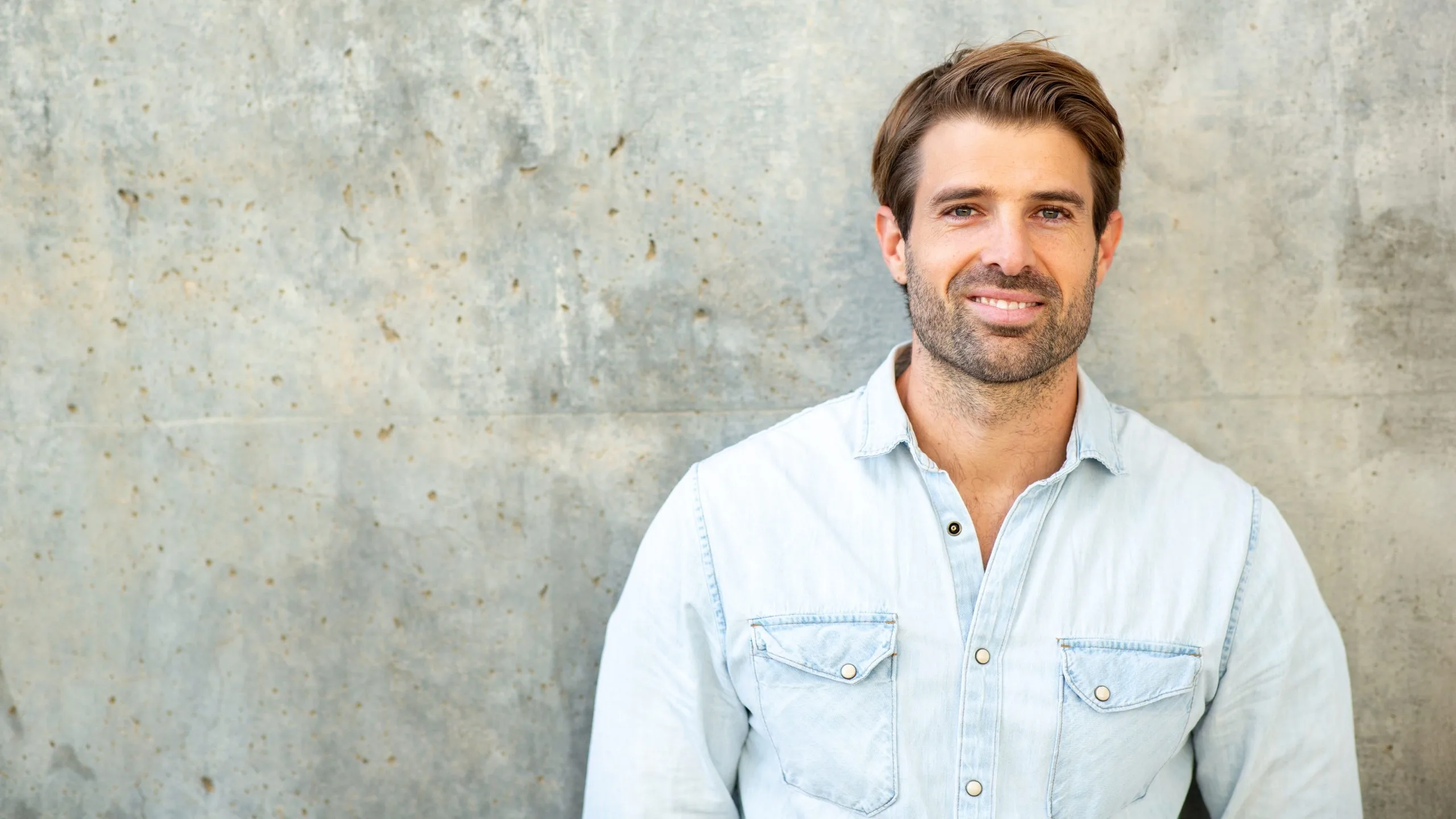 Man in light blue denim shirt smiling against textured concrete wall