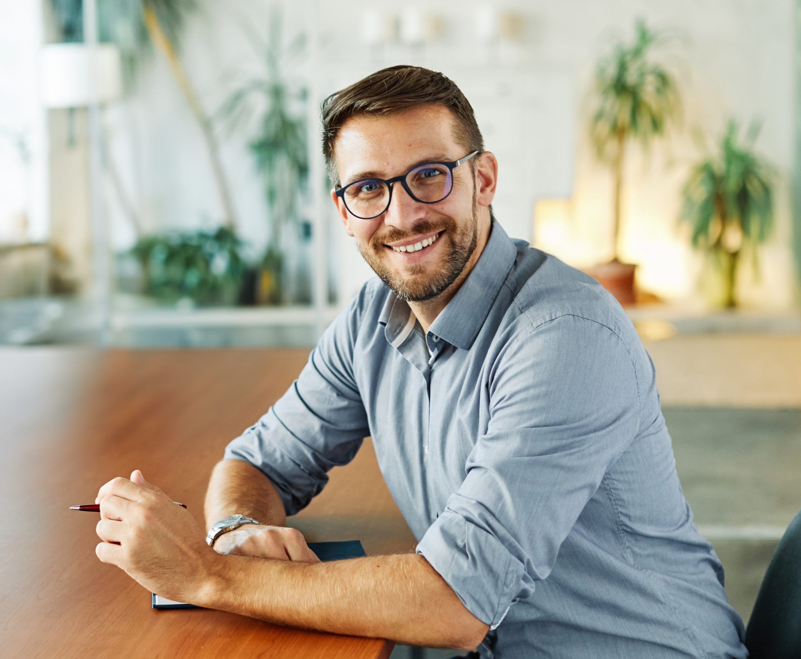 Man in glasses and gray shirt smiling at desk with plants in bright office