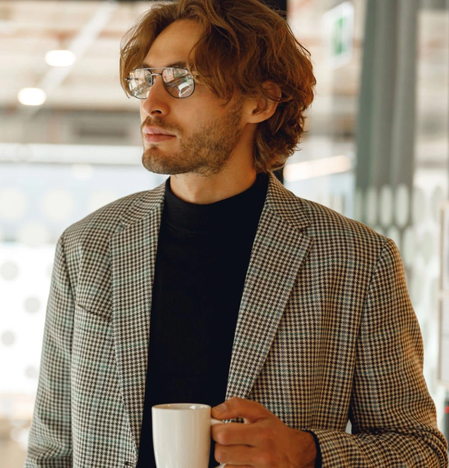 Man wearing glasses and houndstooth blazer holding a coffee mug in a modern office setting
