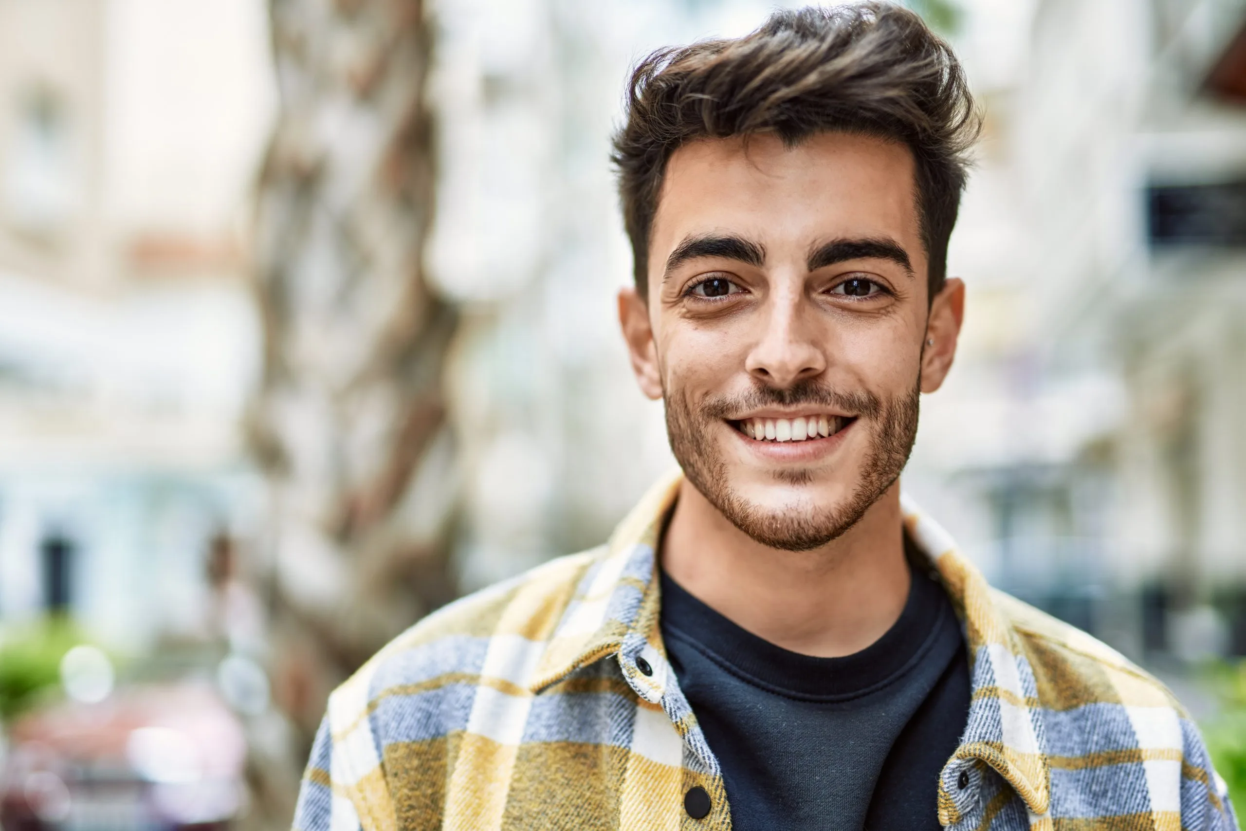 Smiling man in yellow and blue plaid shirt outdoors with blurred urban background