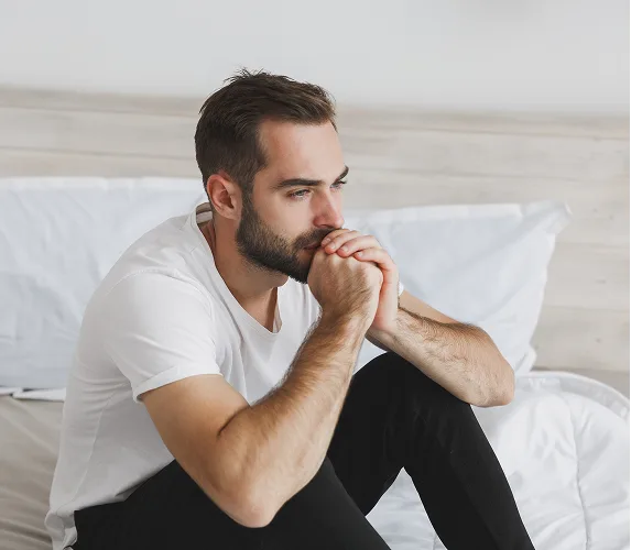 Man sitting on bed with hands clasped, looking away