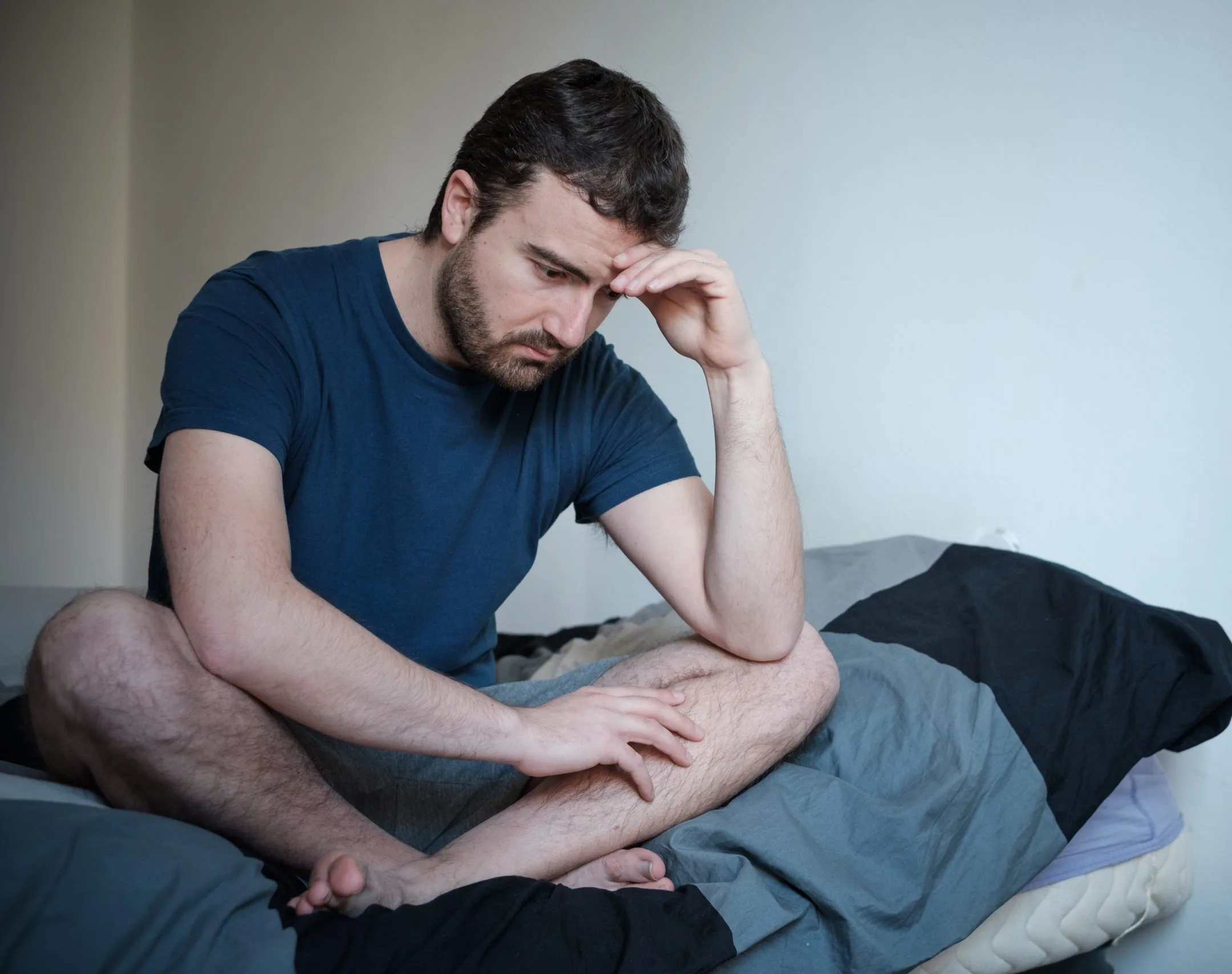 Man sitting on bed with head resting on hand, looking downward with a troubled expression