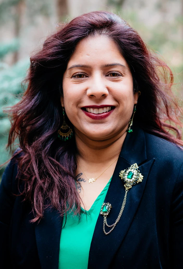 Woman with burgundy wavy hair wearing a dark blazer over a teal top with ornate jewelry, smiling