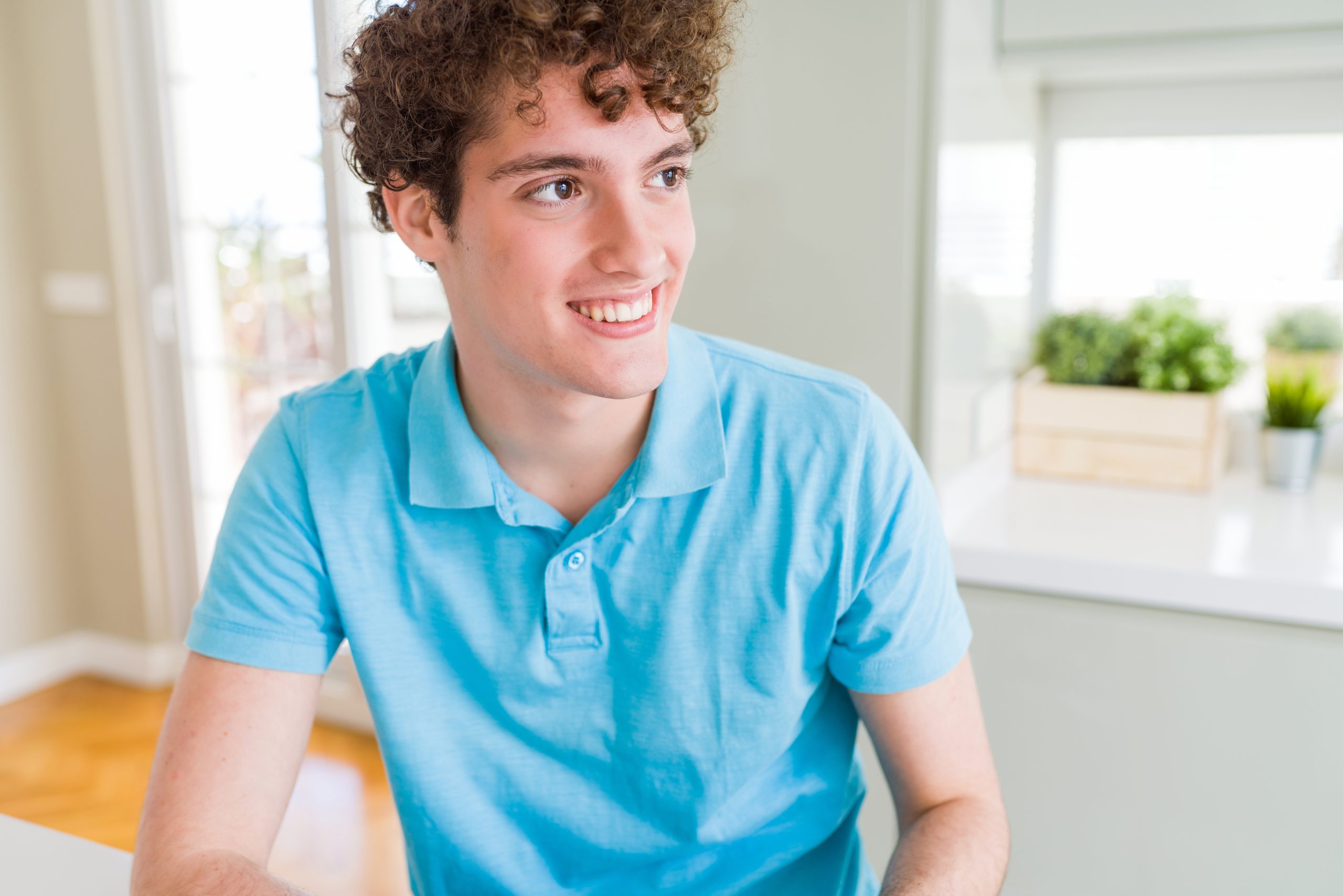 Teenage boy in blue polo shirt smiling by window with plants