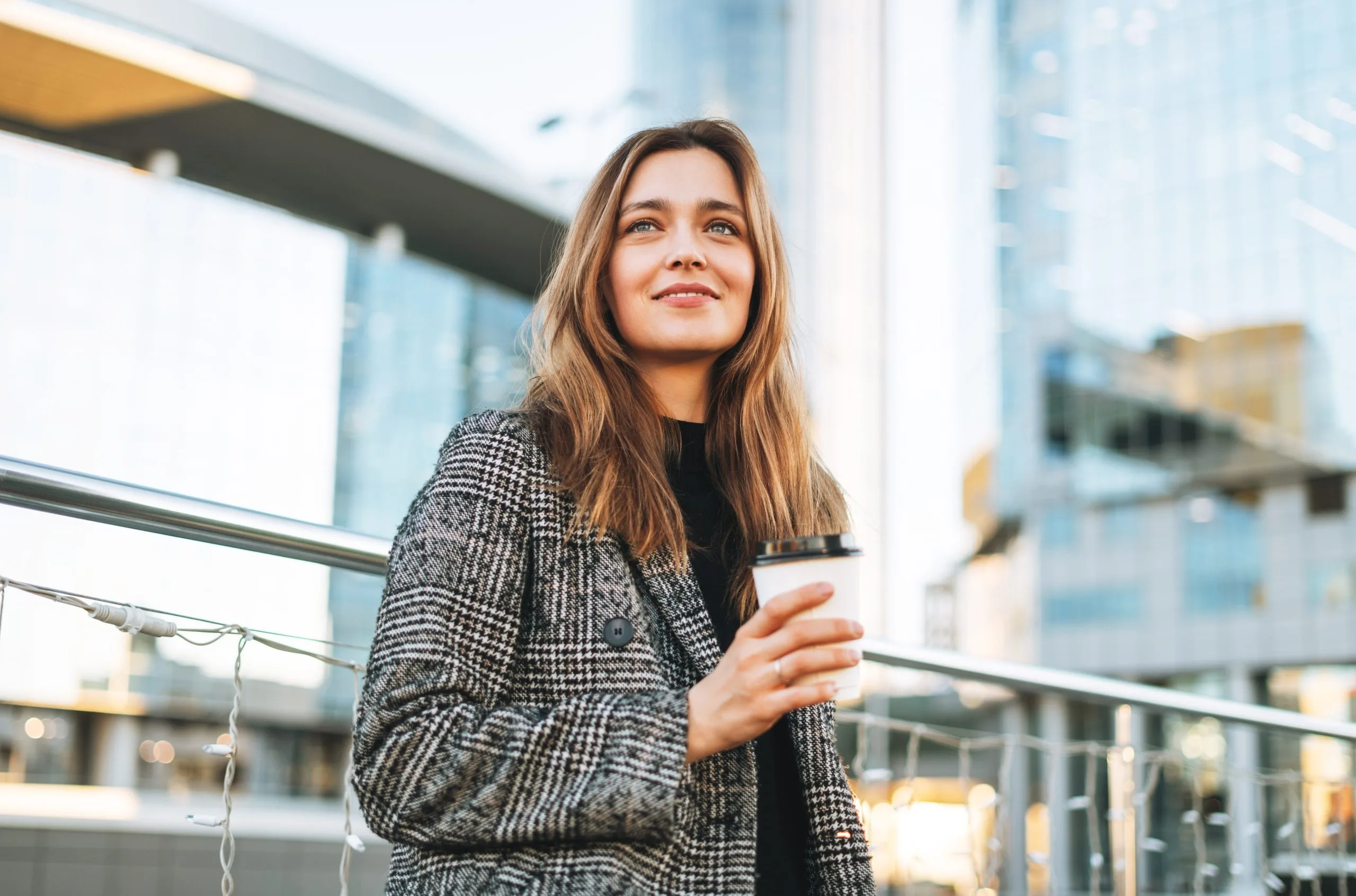 Woman in a patterned blazer holding a coffee cup, smiling outdoors in an urban setting