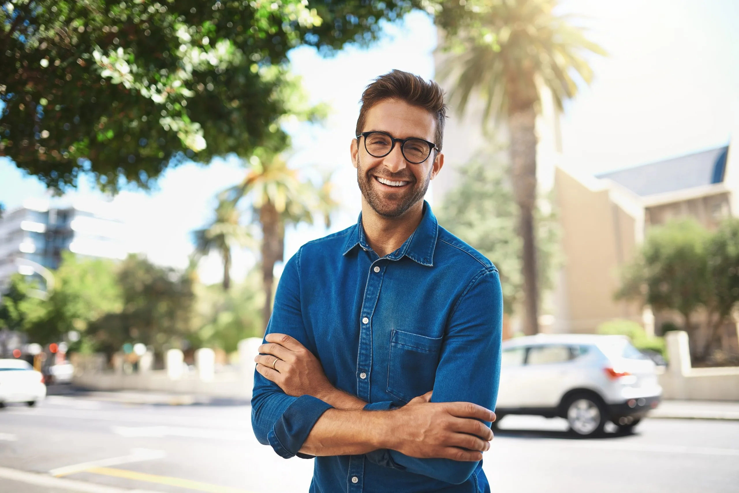 Man in glasses and denim shirt smiling with arms crossed on a sunny city street
