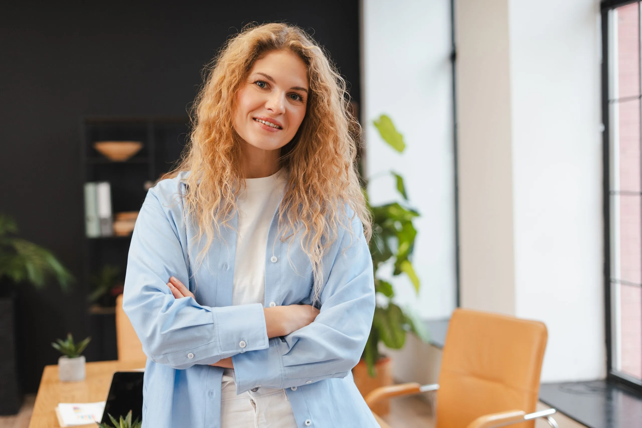 Woman in light blue shirt standing with arms crossed in a modern office with plants and orange chair