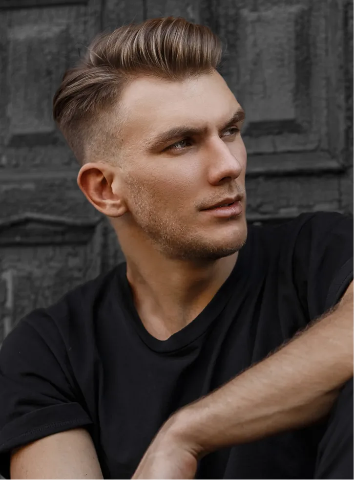 Man with styled dark hair wearing black v-neck shirt against brick wall
