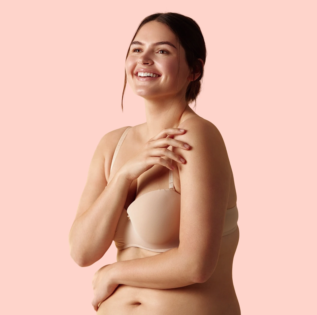 Smiling woman in beige bra against pink background, hands on shoulder and torso