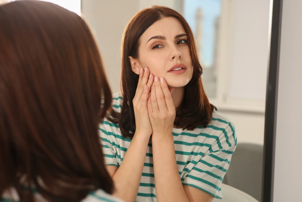 Woman in striped shirt touching her cheek while looking in mirror