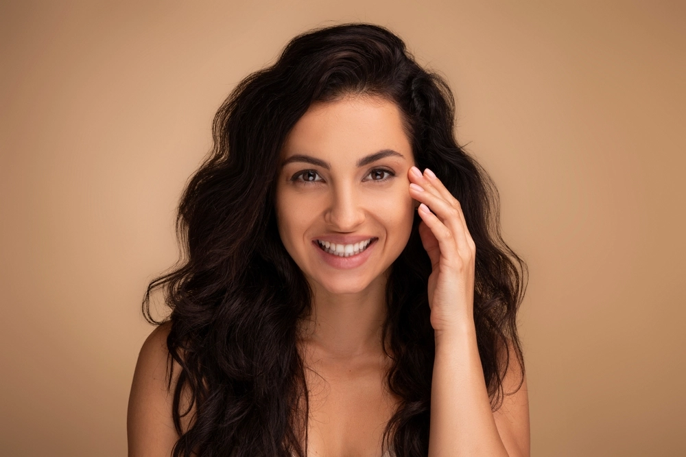 Woman with black wavy hair smiling and touching her face against a beige background