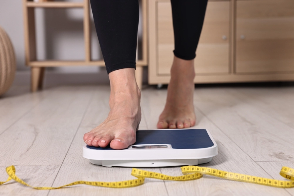 Person stepping onto white bathroom scale with yellow measuring tape nearby