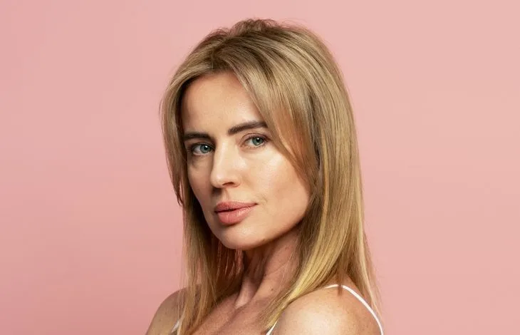 Woman with blonde hair and freckles in white tank top against pink background