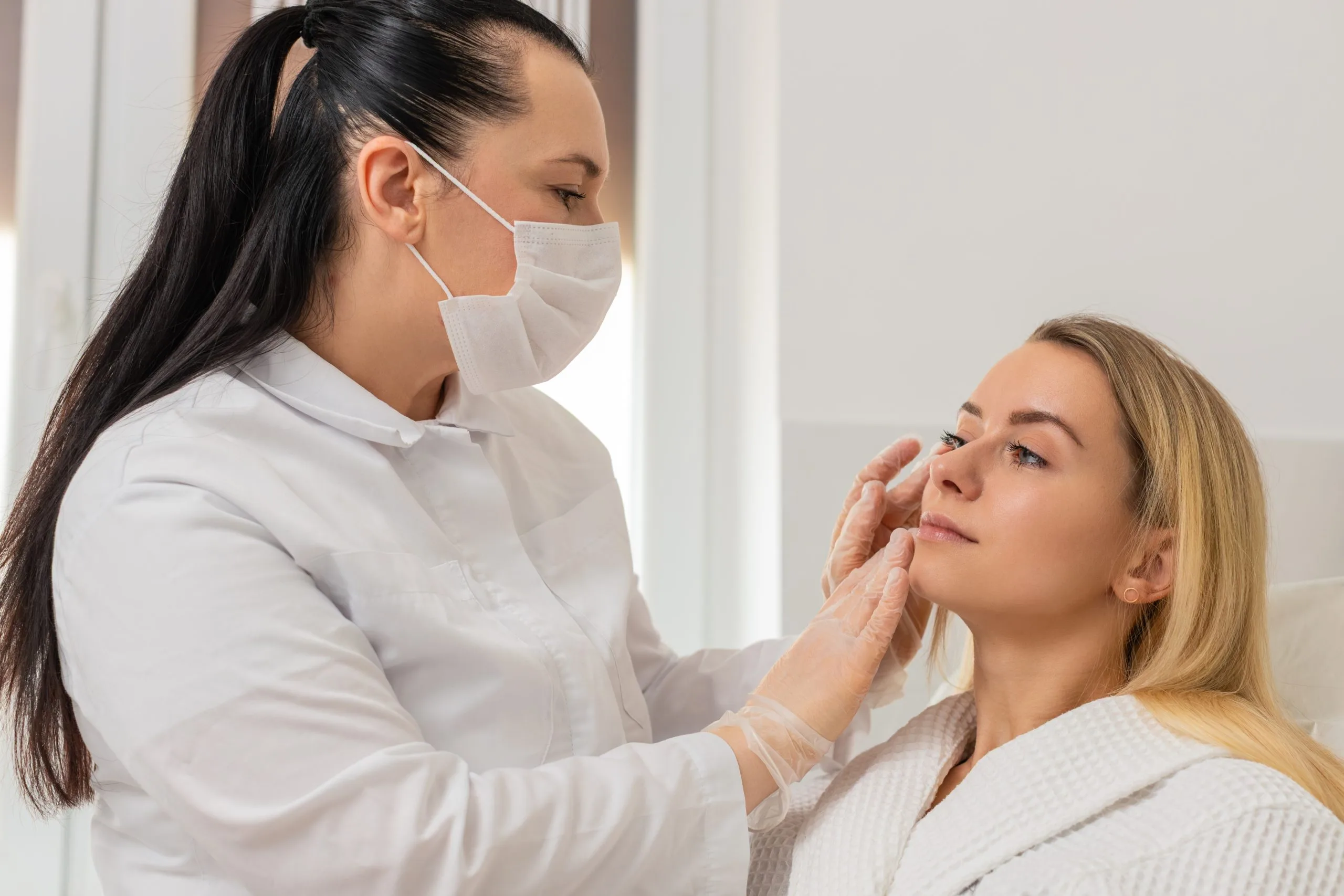 Healthcare provider wearing mask examining patient's face during aesthetic consultation