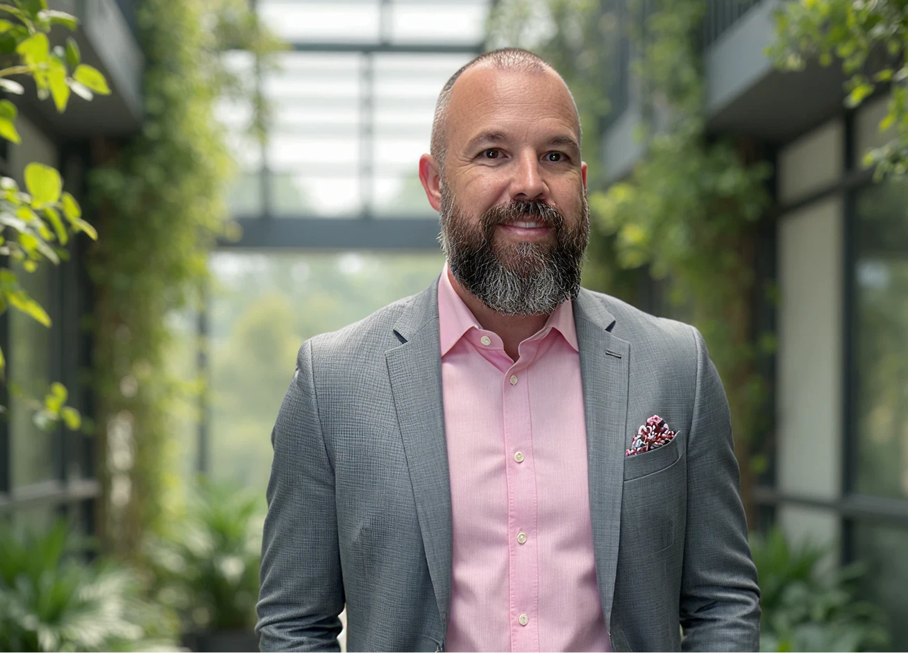 Man in gray blazer and pink shirt standing in a plant-filled corridor