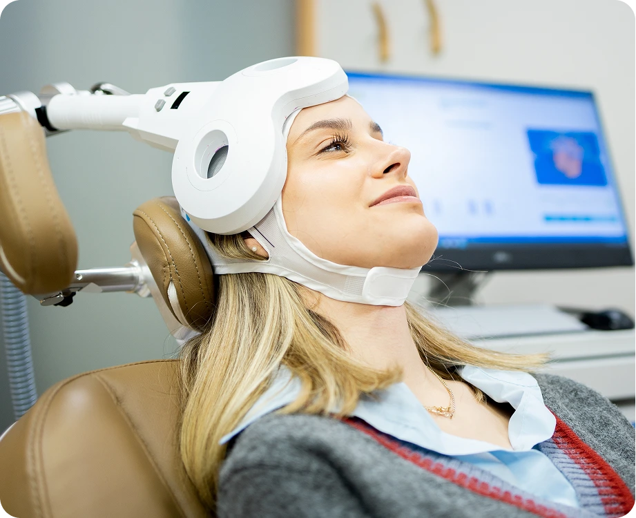 Patient receiving transcranial magnetic stimulation treatment with white headgear while monitoring system displays on screen