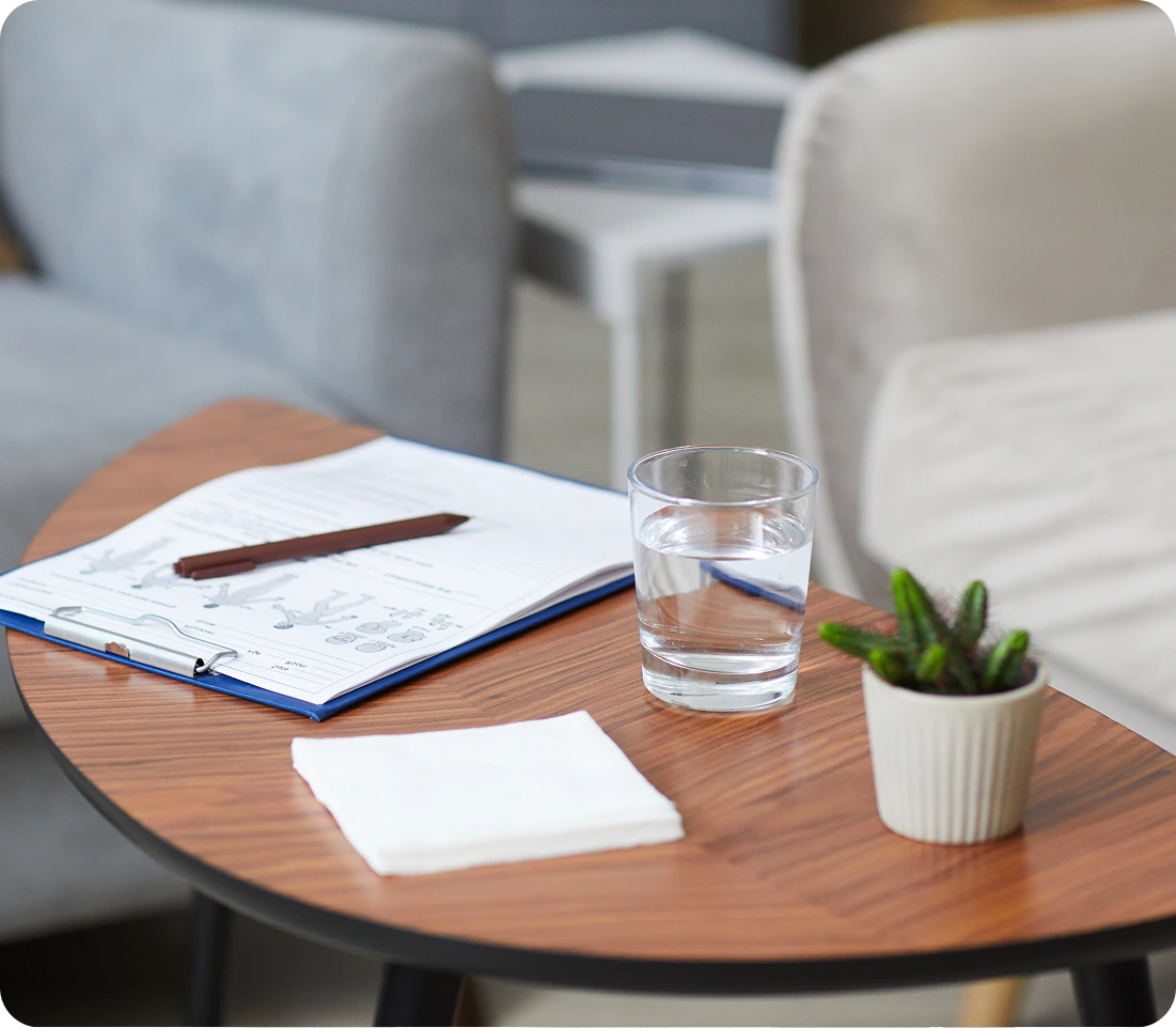 Therapist's desk with open appointment book, pen, glass of water, small potted plant, and tissues in a calm consultation room