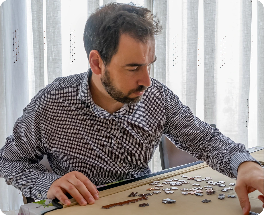 Man in checkered shirt focusing on puzzle pieces on table by window