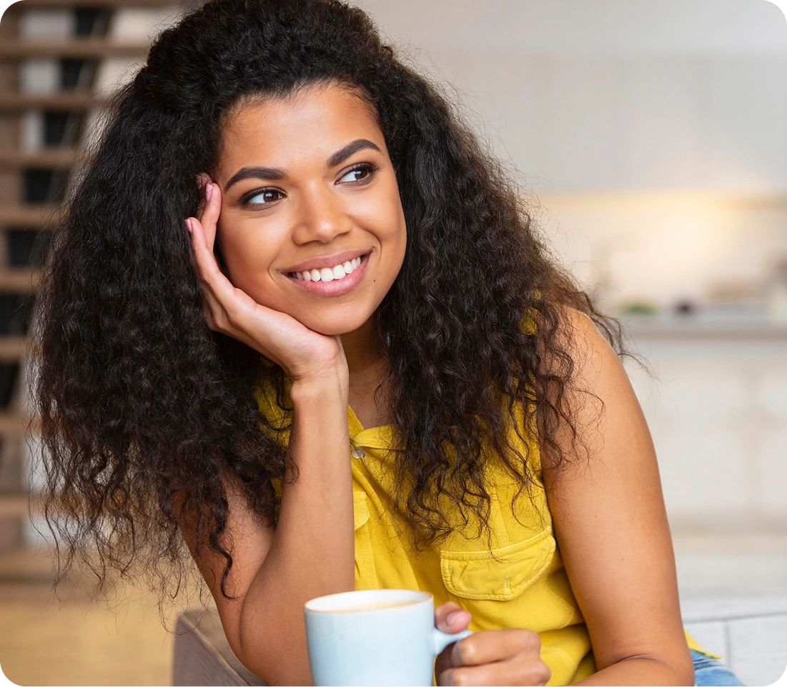 Smiling woman in yellow top holding a mug, hand to face, in a bright modern office setting