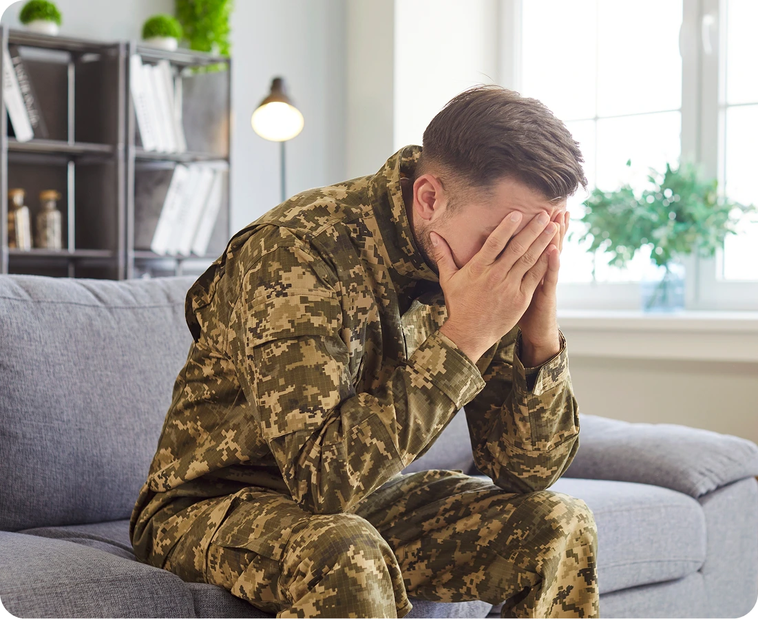 Military service member in camouflage uniform sitting on couch with head in hands, showing distress