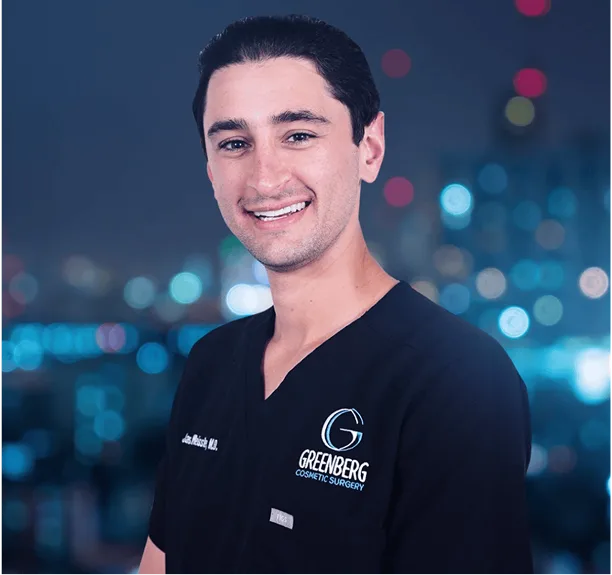 Young man in black Greenberg Cosmetic Surgery scrubs smiling against city lights background