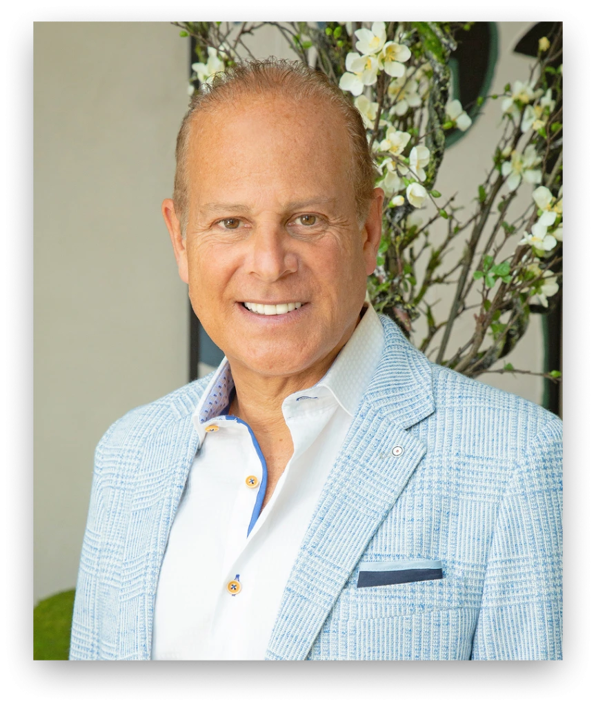 Dr. Stephen T. Greenberg in light blue blazer and white shirt, smiling at camera with white flowers in background