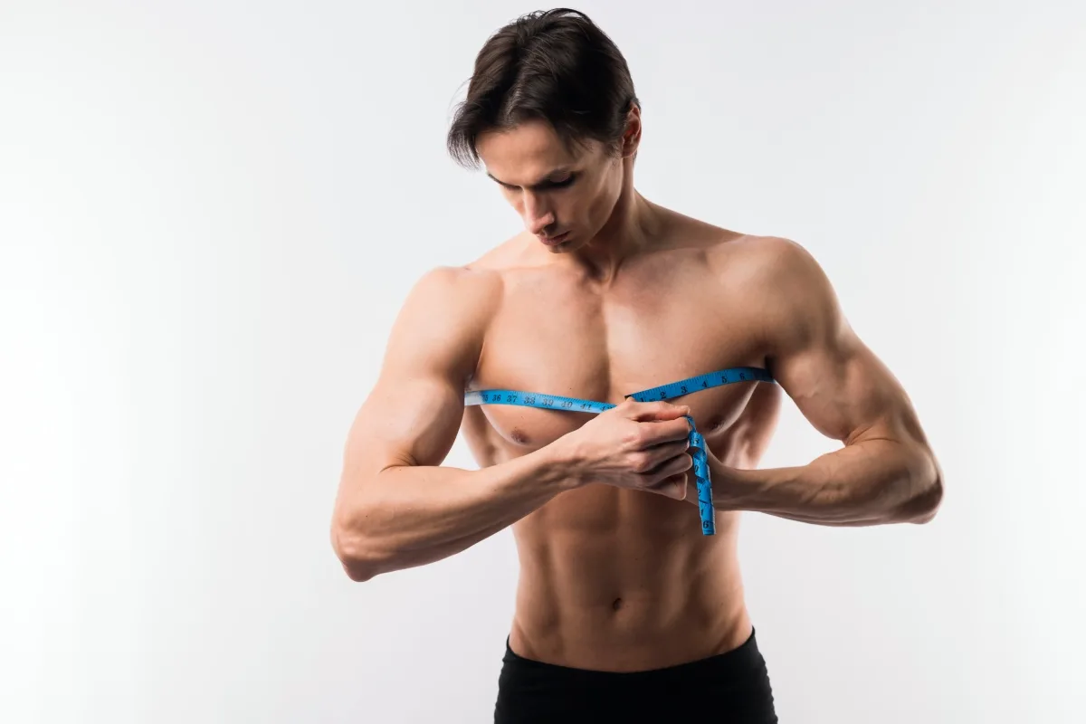 Shirtless man measuring his chest with blue measuring tape against white background