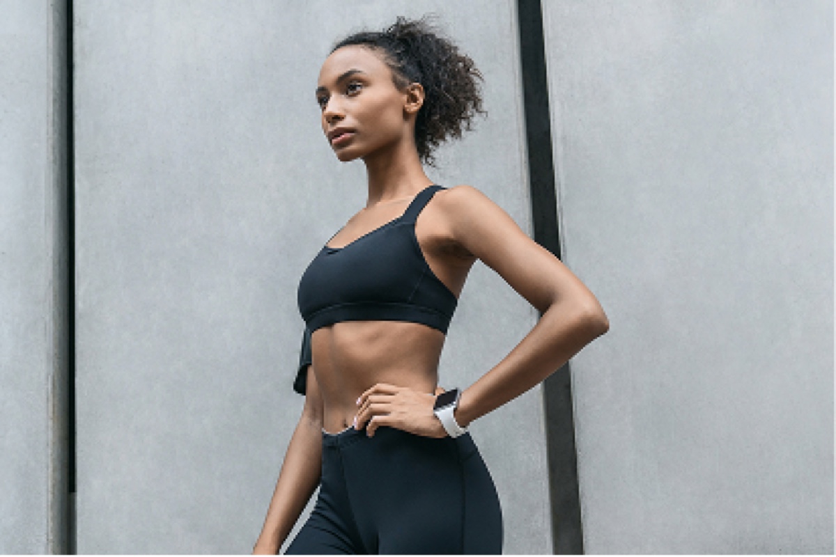 Woman in black sports bra and leggings standing in profile against gray wall