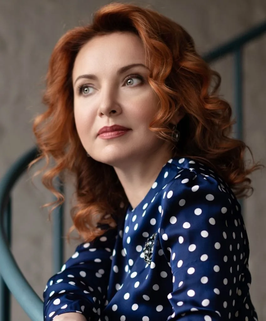 Woman with reddish-brown wavy hair wearing a blue polka-dot top, looking over her shoulder