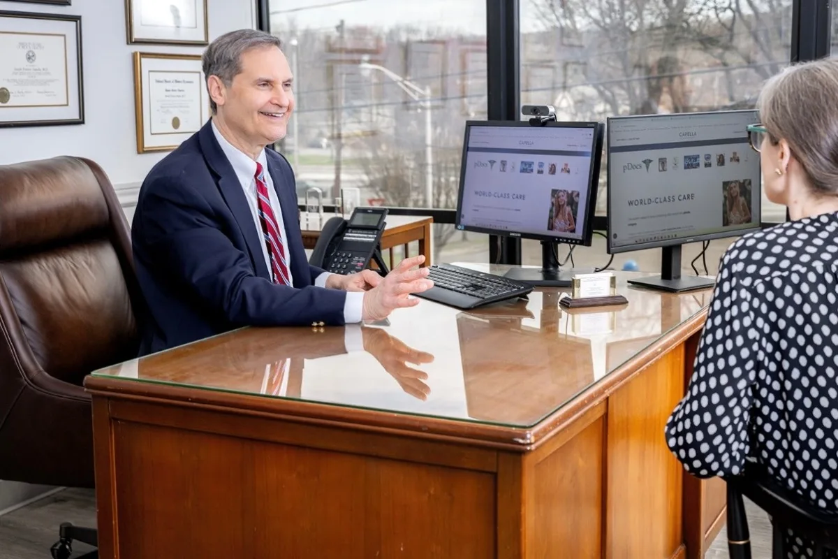 Plastic surgeon in office consulting with patient at desk with dual monitors displaying surgical information