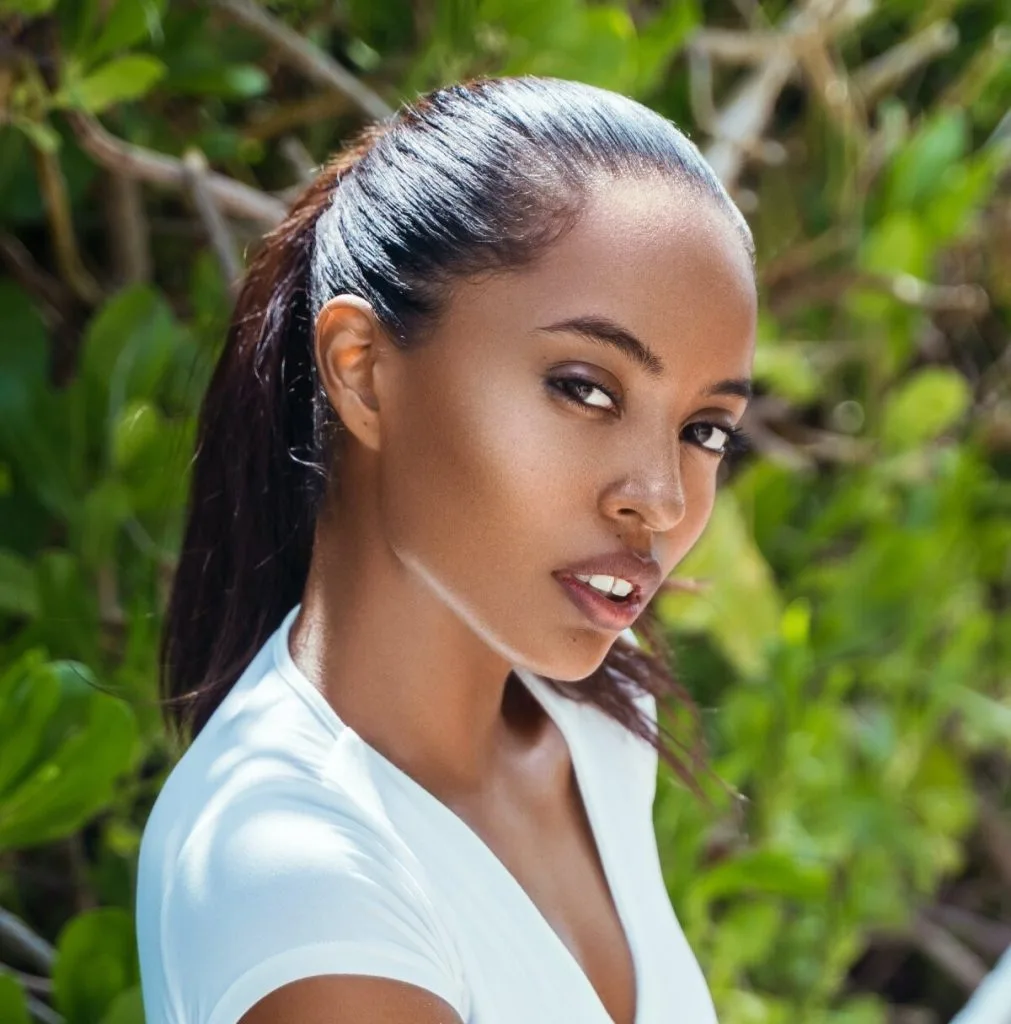 Woman with dark hair in a bun wearing a white top, standing outdoors among green foliage