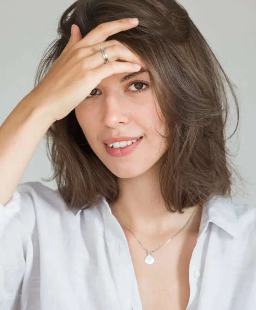 Woman with shoulder-length brown hair smiling while touching her forehead with her hand