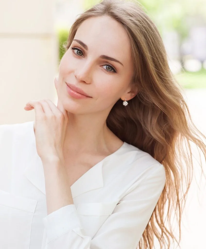 Woman with reddish-brown hair in white top, touching her face and looking at camera with natural outdoor background