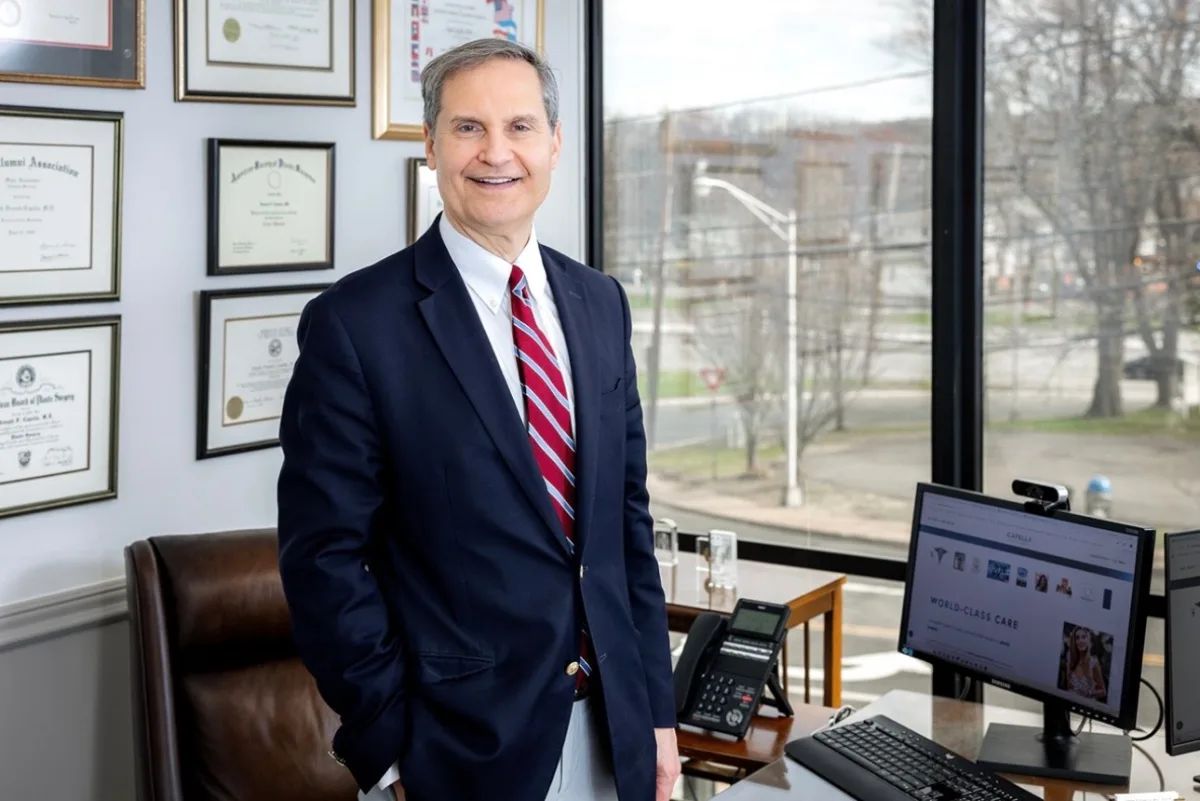Surgeon in dark suit and red striped tie standing in office with diplomas on wall and city view