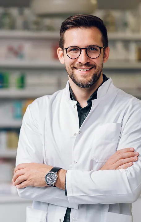Pharmacist in white coat and glasses standing with arms crossed in pharmacy