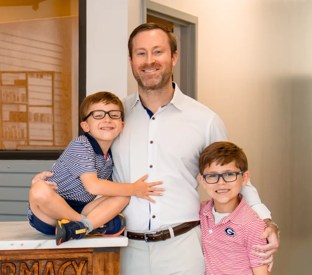 Man in white shirt with two young boys wearing glasses smiling in a pharmacy setting