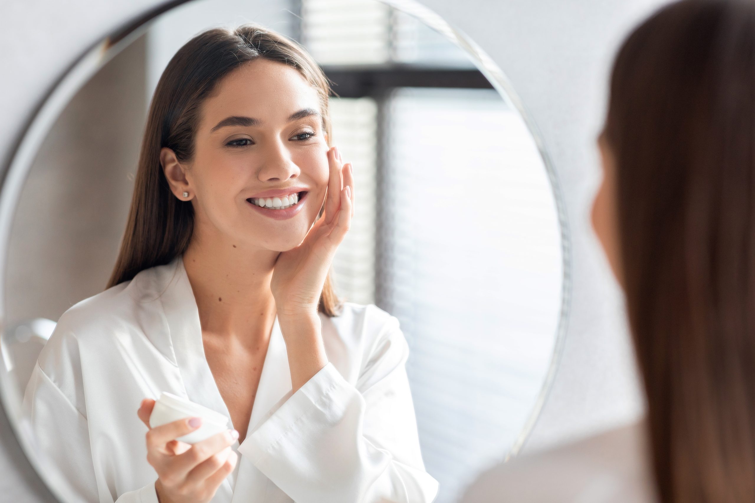 Woman smiling at mirror while applying skincare product to her face