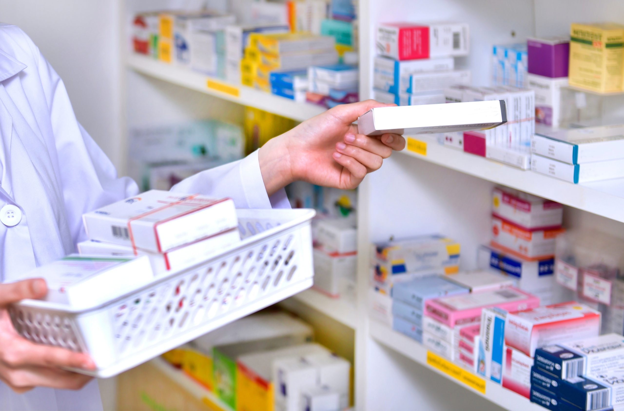 Pharmacist in white coat selecting medication bottle from shelf stocked with colorful drug boxes