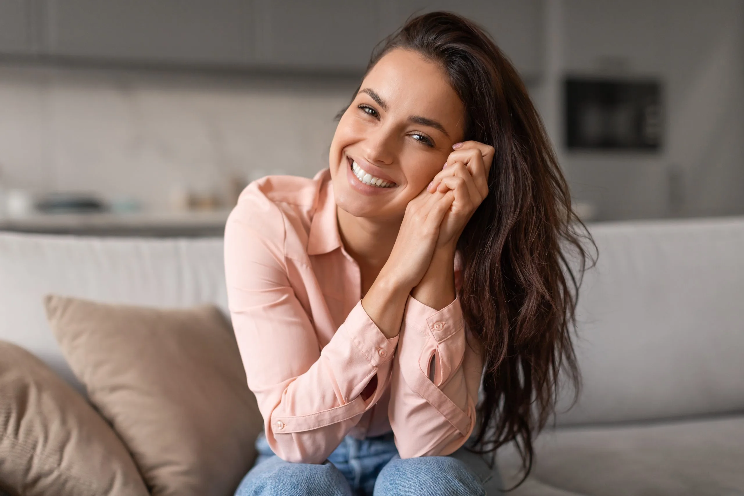Woman in pink shirt smiling on sofa with hand to face