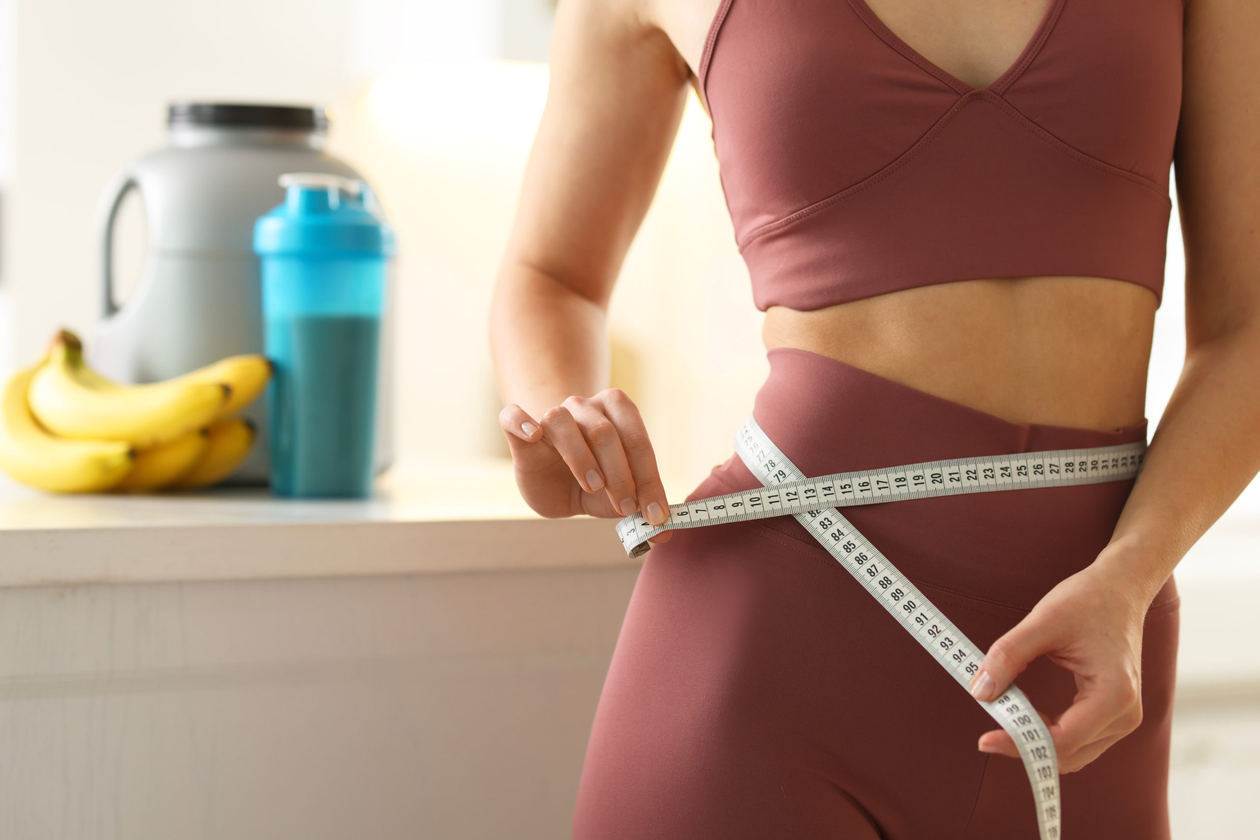 Woman in athletic wear measuring her waist with tape measure near water bottle and bananas