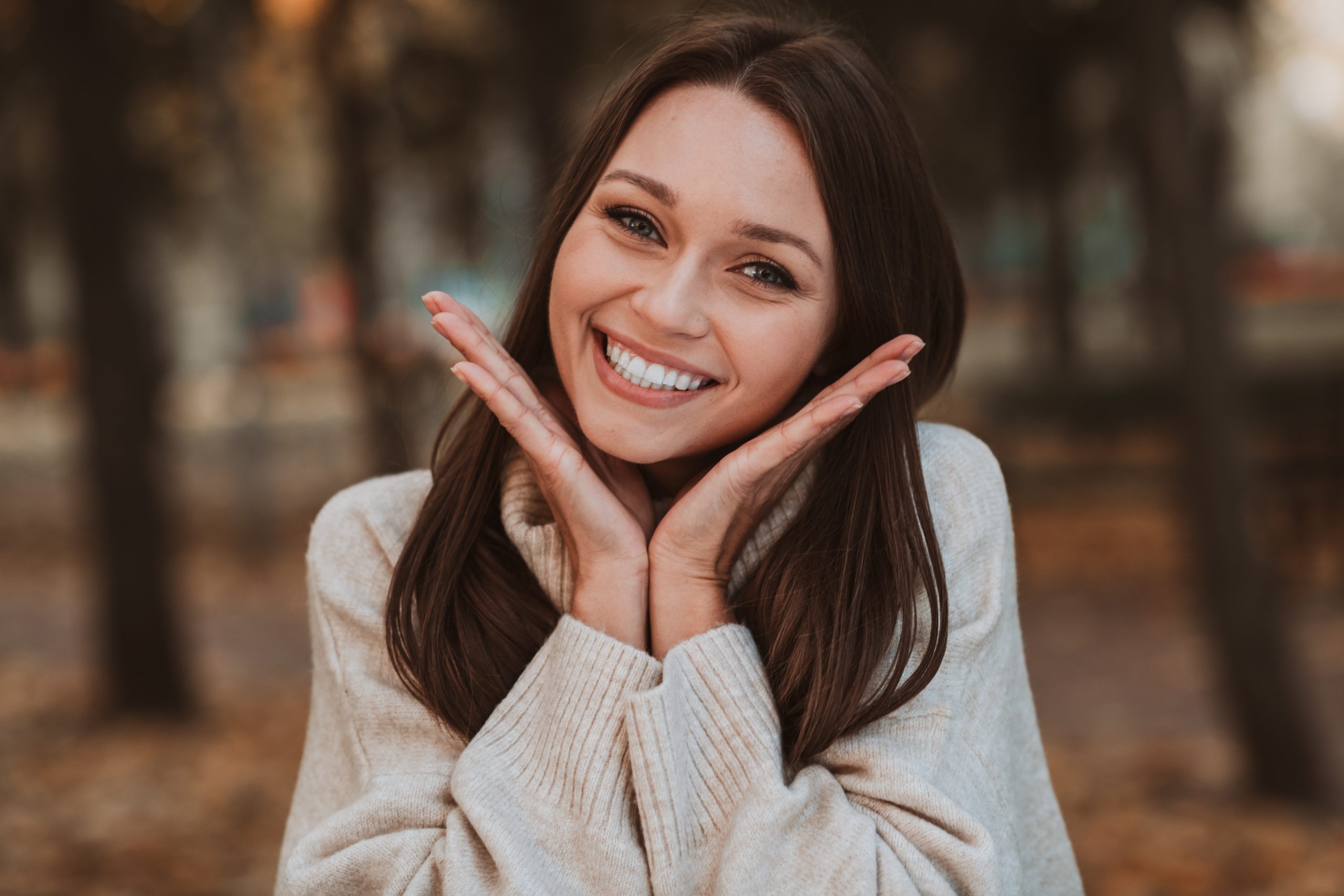 Woman smiling outdoors with hands near face showing healthy complexion
