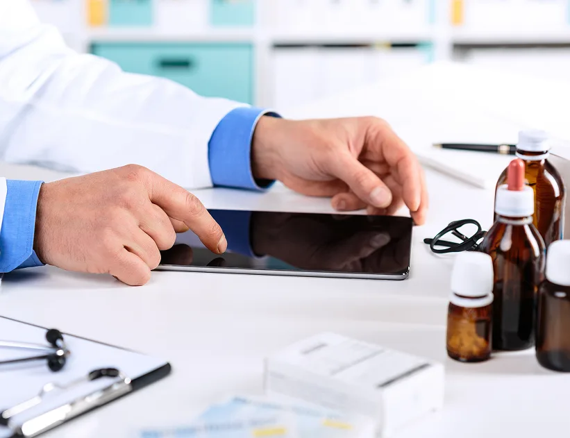 Healthcare provider in white coat with blue cuffs using tablet near medication bottles and medical documents