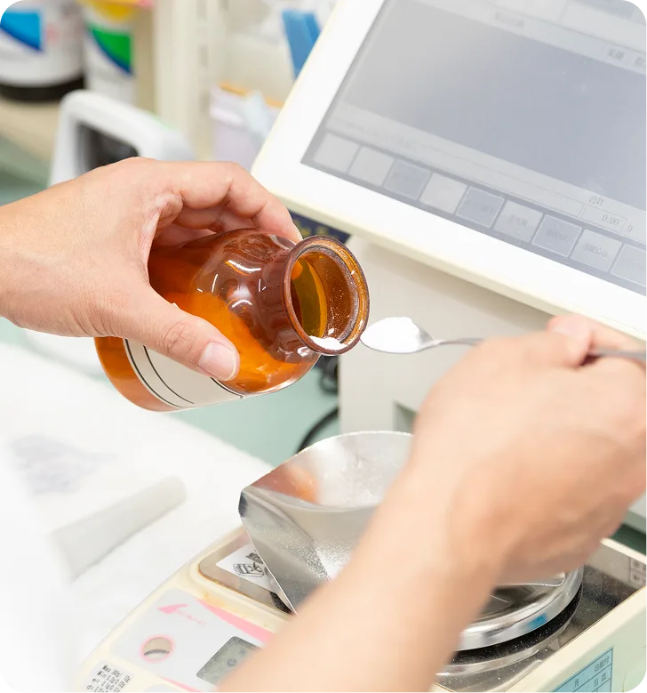 Pharmacist pouring amber liquid from bottle into white bowl at compounding workspace with laptop and materials