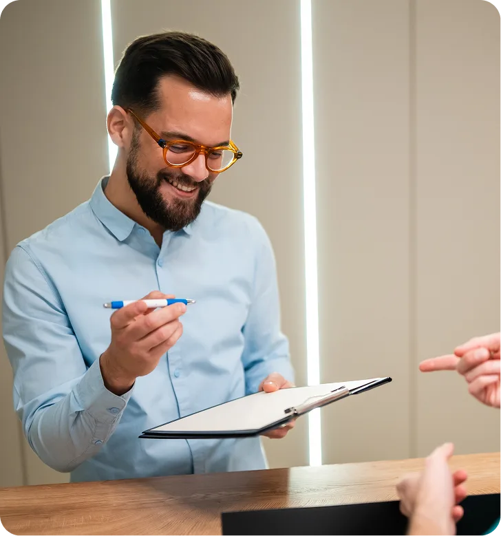 Man in light blue shirt and orange glasses holding pen and clipboard while discussing with another person