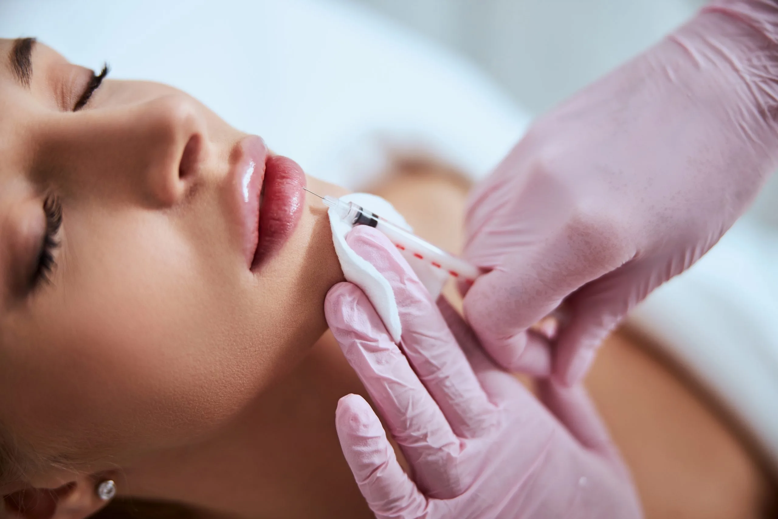 Practitioner in pink gloves applying lip filler injection to reclined patient
