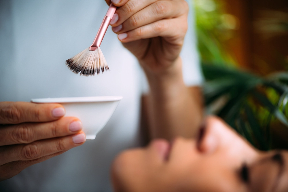 Person holding pink makeup brush above white bowl with cosmetic product
