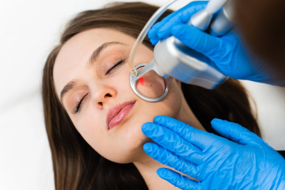 Clinician in blue gloves using a handheld device on a patient's cheek during a facial aesthetic procedure